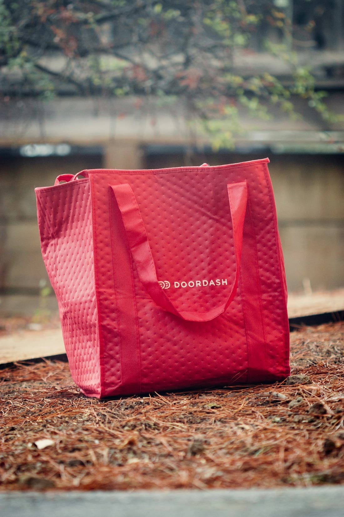 A close-up photograph of a red DoorDash insulated delivery bag sitting on the ground. The bag is textured with a quilted pattern and has two handles. The DoorDash logo is visible in white on the lower part of the bag. The ground is covered with pine needles and small branches, suggesting an outdoor setting. The background is slightly blurred, with a building or wall visible. The lighting is natural, casting a soft shadow behind the bag.