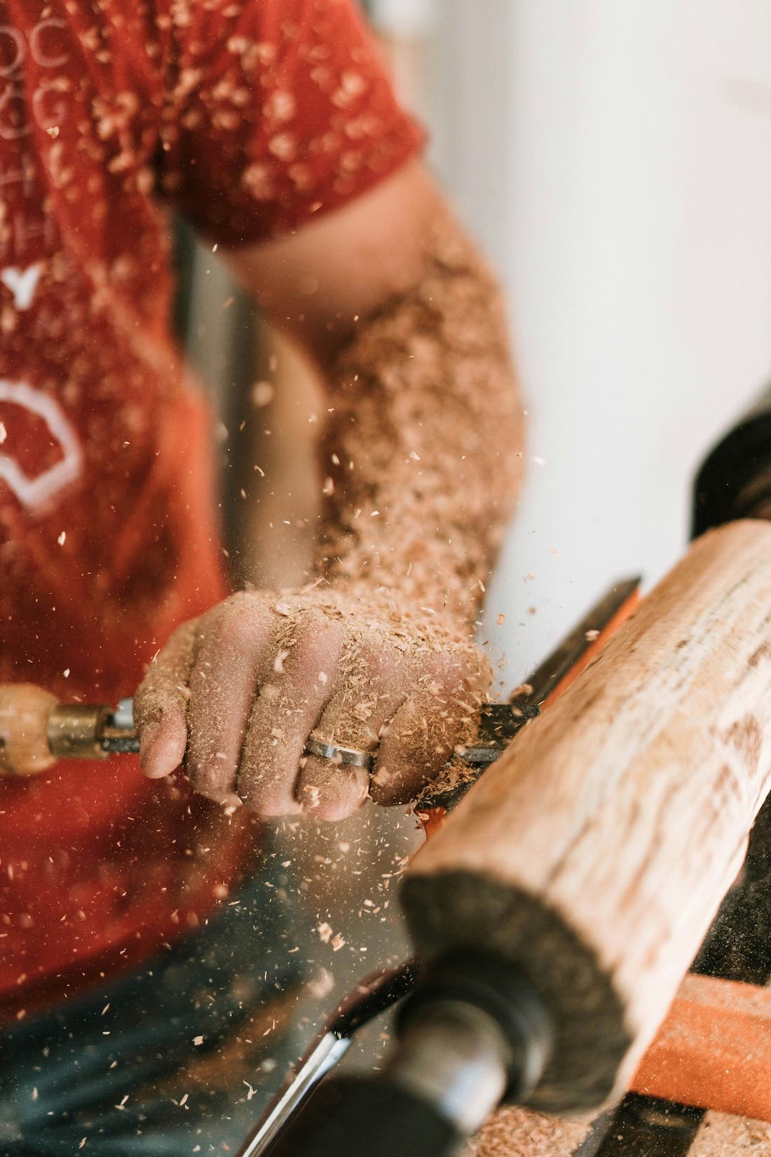 A close-up photograph of a person's hands woodworking. The person is using a tool to shape a piece of wood that is mounted on a lathe. Wood shavings are flying off the wood as it is being worked. The person is wearing a red shirt, and only their hands and forearms are visible.