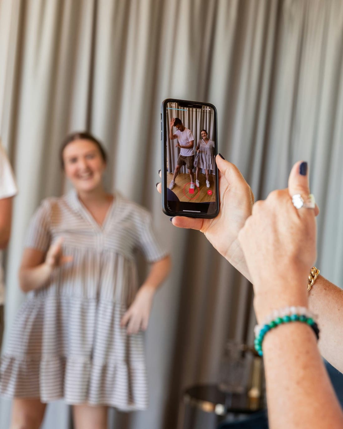A person's hands hold a smartphone, positioned vertically, recording a video. The phone screen shows a man and a woman in light-colored clothing dancing. In the background, slightly out of focus, is a woman in a striped dress, smiling and appearing to be posing or dancing. Light-colored curtains are visible behind her. The person holding the phone has several bracelets on their wrist and rings on their fingers.